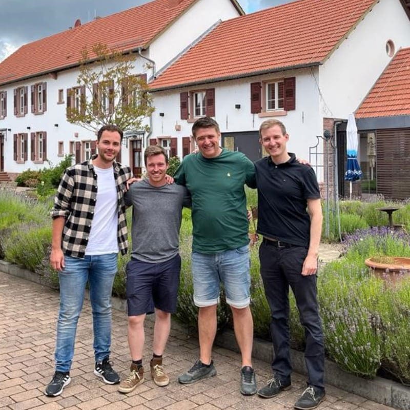 4 Männer in Arbetiskleidung stehen zusammen vor einem Beet vor einer Seite des Hofes.