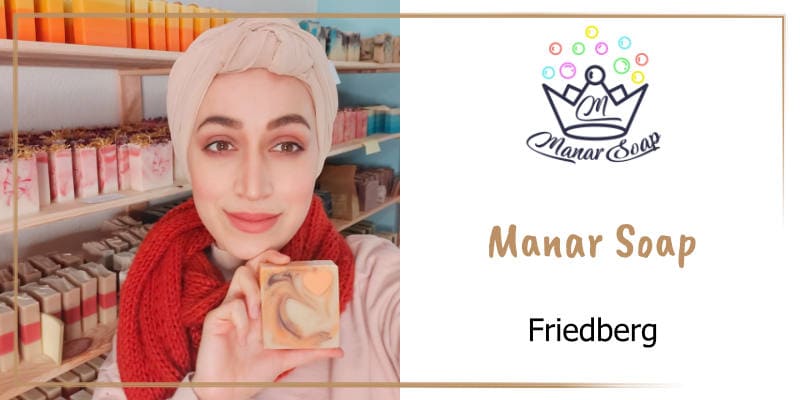 Auf der goldbraun gerahmten WasRegionales-Markenkachel Manar Alkhatib freundlich laechelnd zeigt ein Stueck Seife, im Hintergrund ihre bunt gefuellten Seifenregale; das Logo von Manar Soap mit der Krone und Seifenblasen; darunter der Schriftzug Manar Soap