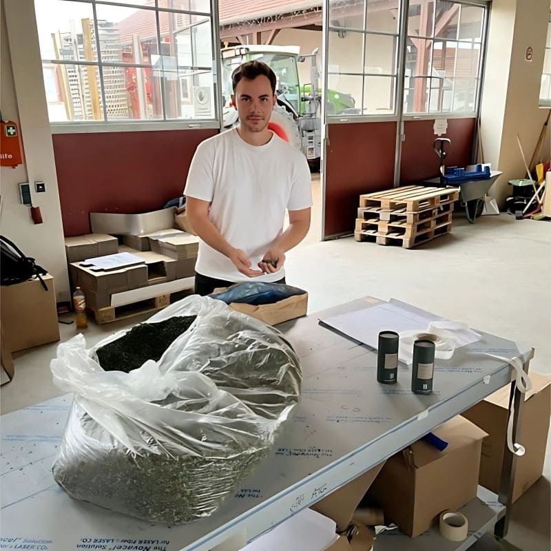 junge rMann an Arbeitstischin Arbeitshalle. Auf dem Tisch ein SAck mit getrocknetem Kräutertee, Kräutertee-Pappdosen und eine Waage