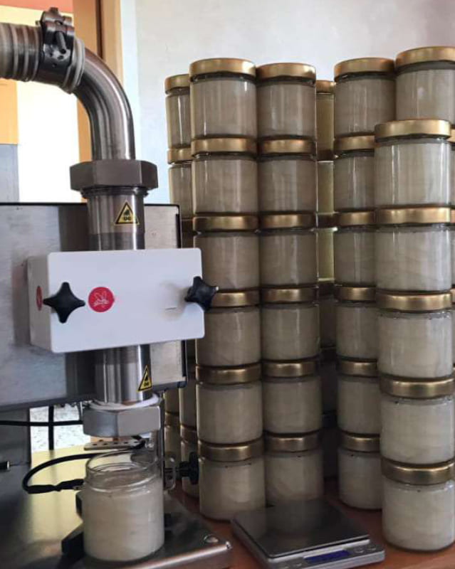 LInksseitig die Abfüllmaschine mit darunter stehendem gefüllten Honigglas. Rechts viele gestapelte Honiggläser